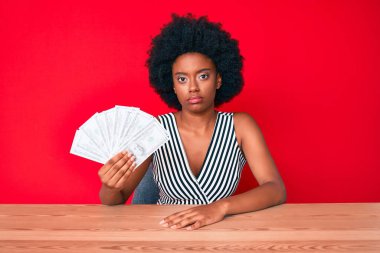 Genç Afrikalı Amerikalı kadın dolarları tutuyor. Düşünen tutum ve ayık yüz ifadesi. Kendinden emin görünüyor. 
