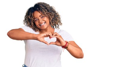 Genç Afro-Amerikan artı cüsseli kadın, gündelik kıyafetler giyiyor, aşık gülümsüyor, elleriyle kalp sembolü yapıyor. romantik kavram. 