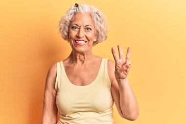 Kıdemli, gri saçlı, günlük kıyafetler giyen, üç numaralı parmaklarıyla işaret eden, kendinden emin ve mutlu bir şekilde gülümseyen kadın.. 