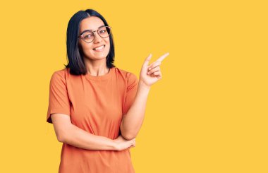 Yüzünde kocaman bir gülümseme olan, gündelik kıyafetler giyen genç ve güzel Latin bir kadın, kameraya el ve parmak ile bakıyor.. 