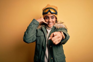 Genç Afrikalı Afro-Amerikan kayakçı kız kar maskesi ve kayak gözlüğü takıyor gülümsüyor telefonda konuşuyor ve seni işaret ediyor. Ara beni..