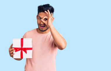 Yakışıklı Latin Amerikalı genç adam elinde hediyeyle gülümsüyor mutlu bir şekilde el pençe divan duruyor. 