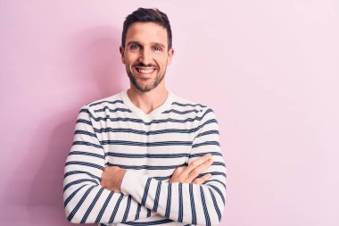 Genç, çizgili süveter giyen yakışıklı bir adam izole edilmiş pembe arka planda durup kollarını kavuşturup kameraya bakarak gülümsüyor. Pozitif kişi.
