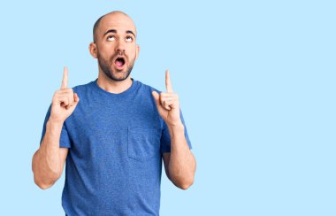 Gündelik tişört giyen genç yakışıklı şaşırmış ve şaşırmış bir şekilde yukarı bakıyor ve parmaklarıyla işaret ediyor ve kollarını kaldırıyor.. 