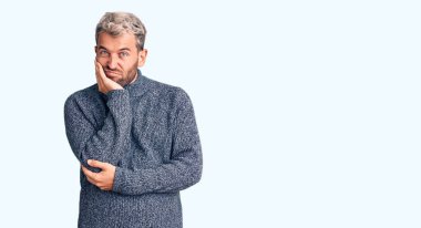 Genç, sarışın, sıradan bir süveter giyen, yorgun ve bunalımdan sıkılmış görünen, kollarını kavuşturmuş genç bir adam.. 