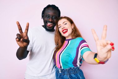 İki elini de zafer işareti yaparak parmaklarını göstererek gülümseyen gündelik kıyafetler giyen ırklar arası bir çift. İki numara.. 