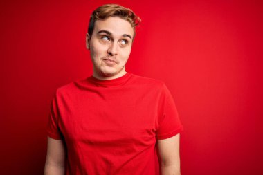 Genç, yakışıklı, kırmızı tişörtlü bir adam izole edilmiş kırmızı arka planda gülümsüyor ve başka yöne bakıyor..
