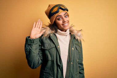 Genç Afrikalı Afro-Amerikan kayakçı kız kar sporu giyer ve kar gözlüğü takar Mutlu ve güler yüzlü hoş geldin jestleri yapar