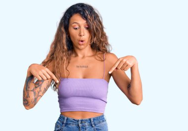 Dövmeli genç İspanyol kadın, günlük giysiler giymiş, parmaklarını göstererek reklamını yapıyor, şaşırmış yüzünü ve ağzını açıyor. 