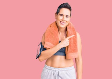 Kısa saçlı, spor kıyafetleri ve havlusu olan genç bir kadın. Akıllı telefon kullanıyor. Yüzünde mutlu ve doğal bir ifade olan el ve parmak ile yüzünü gösteren bir gülümseme var. 