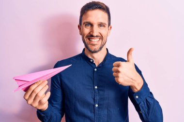 Arkaplanda pembe kağıt uçak tutan genç yakışıklı adam mutlu ve pozitif gülümsüyor, başparmağıyla mükemmel ve onay işareti yapıyor.