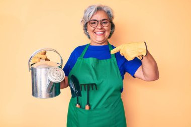 Bahçıvan önlüğü giyen, elinde su dolu eldiven tutan yaşlı, beyaz saçlı kadın gülümseyen mutlu ve gururlu birini işaret ediyor. 