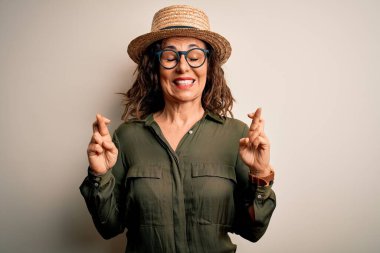 Orta yaşlı esmer kadın, gözlüklü ve şapkalı, izole edilmiş beyaz arka planda duruyor. Umut ve gözler kapalı bir şekilde gülümsüyor. Şans ve batıl inanç..
