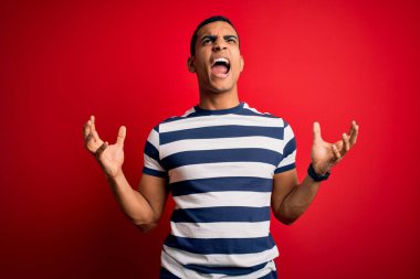 Yakışıklı Afro-Amerikan erkeği, sıradan çizgili tişört giymiş kırmızı arka planda çılgın deli bağırıyor ve agresif bir ifade ve kollarla bağırıyor. Hayal kırıklığı konsepti.