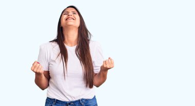 Genç ve güzel esmer kadın, gündelik tişört giyip mutlu ve heyecanlı bir şekilde kolları havada, gülümseyerek ve başarı için bağırarak kazanan jestler yapıyor. kutlama konsepti. 