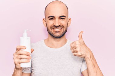 Genç yakışıklı kel adam elinde krem losyon şişesiyle mutlu ve pozitif gülümsüyor, başparmağıyla mükemmel ve onay işareti yapıyor. 