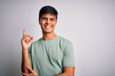 İzole edilmiş beyaz arka plan üzerinde duran sıradan bir tişört giyen genç ve yakışıklı bir adam. Yüzünde kocaman bir gülümseme, eli ve parmağı kameraya bakıyor..