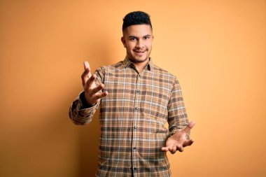 Sarı arka plan üzerinde duran sıradan tişörtlü yakışıklı genç adam güler yüzlü, güler yüzlü, dost canlısı, olumlu ve kendinden emin selamlar.