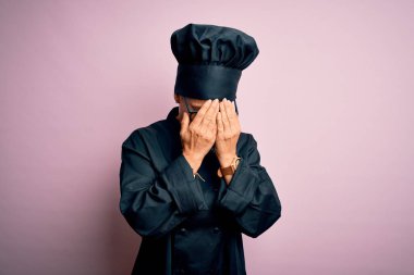 Orta yaşlı esmer aşçı kadın aşçı üniforması giyiyor ve pembe arka planda şapkası var. Üzücü bir yüz ifadesi var. Ağlarken yüzünü kapatıyor. Depresyon kavramı.