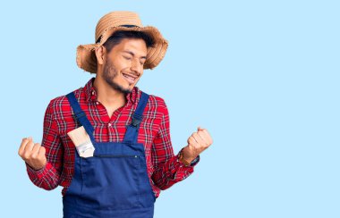 Yakışıklı Latin Amerikalı, tamirci üniforması giyen çok mutlu ve heyecanlı bir genç. Kolları havada, gülümseyen ve başarı için bağıran kazanan bir jest yapıyor. kutlama konsepti. 