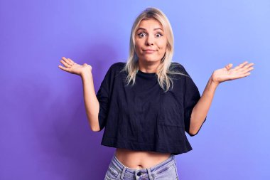 Siyah bir tişört giyen genç ve güzel bir sarışın. Mor arka planda duruyor. Kolları açık, hiçbir fikri yok ve şüpheli bir yüzü var..