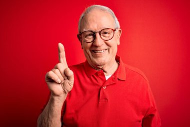 Gri saçlı, gözlüklü, kırmızı arka planda rahat tişörtlü, kendinden emin ve mutlu bir şekilde gülümseyen bir numaralı parmağı gösteren son sınıf öğrencisi..