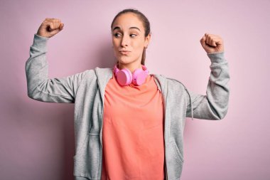 Spor yapan güzel bir kadın müzik dinliyor pembe arka planda kulaklık takıyor ve gururla gülümseyen kol kaslarını gösteriyor. Fitness konsepti.