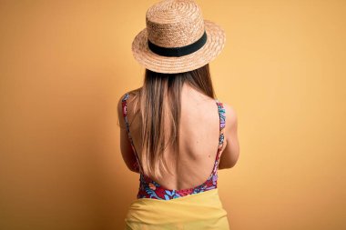 Sarı arka planda mayo ve yaz şapkası takan sarışın güzel bir kadın kollarını kavuşturup arkana bakıyor.