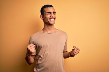 Sarı arka plan üzerinde sıradan tişörtler giyen genç ve yakışıklı Afro-Amerikan erkeği çok mutlu ve heyecanlı bir şekilde kollarını kaldırıp, gülümseyerek ve başarı için çığlık atarak zafer hareketi yapıyor. Kutlama konsepti.
