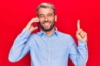 Sakallı, genç, yakışıklı sarışın bir adam akıllı telefondan konuşuyor. Bir fikir veya soru işareti ile gülümsüyor.