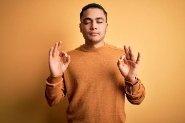İzole edilmiş sarı arka planda dikilen, rahat ve gülümseyen Brezilyalı genç adam parmaklarıyla meditasyon hareketi yapıyor. Yoga konsepti.