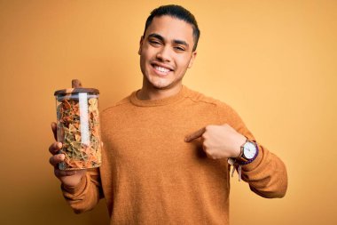 İzole edilmiş sarı arka planda İtalyan makarnası ile dolu kavanozu tutan genç Brezilyalı bir adam kendini işaret eden sürpriz bir yüzle.