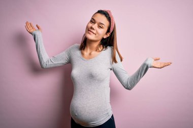 Genç, güzel bir kız hamile, pembe arka plan üzerinde bebek bekliyor. Kolları ve elleri havada, kafası karışmış bir ifade. Şüphe kavramı.