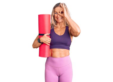 Orta yaşlı, zinde, yoga minderi tutan, gülümseyen, mutlu, el pençe divan duran sarışın bir kadın. 