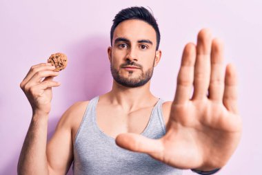 Sakallı, kolsuz tişört giyen, eli açık çikolatalı kurabiye yiyen, ciddi ve kendinden emin bir ifadeyle dur işareti yapan, savunma jesti yapan yakışıklı bir genç.