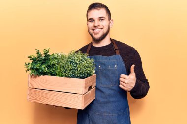 Bahçıvan önlüğü giymiş genç yakışıklı adam elinde pantolon çanağı, gülümsüyor mutlu ve pozitif, başparmağıyla mükemmel ve onay işareti yapıyor. 