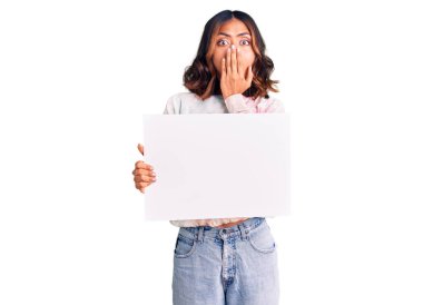 Genç, güzel, melez bir kadın elinde karton pankart, ağzı boş, ağzı elle kaplanmış, şok olmuş ve hata yapmaktan korkan bir kadın. şaşırmış ifade 