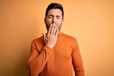 Sarı arka planda dikilen sıradan süveter giyen sakallı yakışıklı genç adam ağzıyla esnemekten sıkılmış. Huzursuz ve uykulu..