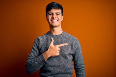 İzole edilmiş turuncu arka planda dikilen sıradan bir süveter giyen genç ve yakışıklı bir adam. Yüzünde mutlu ve doğal bir ifadeyle el ve parmak uçlarını gösteren bir gülümseme.