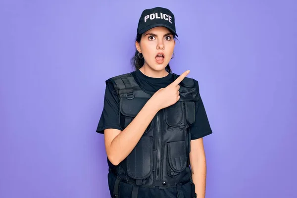 Young police woman wearing security bulletproof vest uniform over purple background Surprised pointing with finger to the side, open mouth amazed expression.