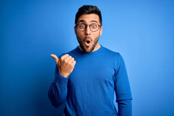 Young handsome man with beard wearing casual sweater and glasses over blue background Surprised pointing with hand finger to the side, open mouth amazed expression.
