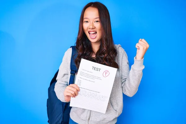 Young beautiful chinese girl showing a failed exam screaming proud, celebrating victory and success very excited with raised arms