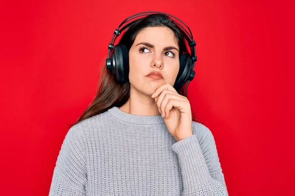 質問について考える顎の手で音楽を赤の背景に耳を傾け 現代のヘッドフォンを身に着けている若い美しい女の子 集中的な表現 思慮深い顔で微笑んだ 疑いの概念 — ストック写真
