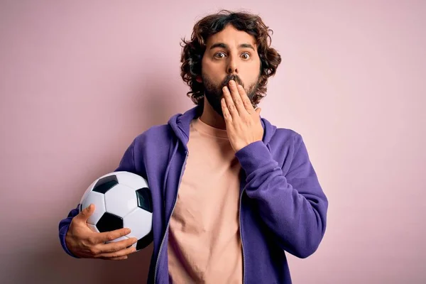 Handsome player man with beard playing soccer holding football ball over pink background cover mouth with hand shocked with shame for mistake, expression of fear, scared in silence, secret concept