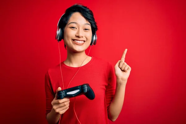 年轻美丽的亚裔游戏玩家女孩用操纵杆和耳机玩电子游戏时 会惊讶地想到一个主意或问题 那就是用快乐的脸蛋指手画脚 位居第一 — 图库照片