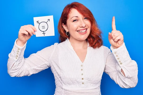 Redhead woman asking for sex discrimination holding paper with transgender equality message smiling with an idea or question pointing finger with happy face, number one