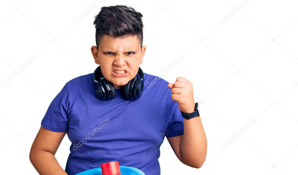 Little boy kid holding skate and wearing headphones annoyed and frustrated shouting with anger, yelling crazy with anger and hand raised 