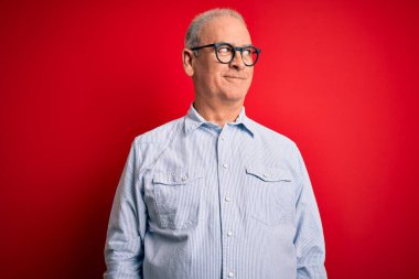 Orta yaşlı, yakışıklı, beyaz, sıradan çizgili tişört giyen, kırmızı arka planda gözlüklü, gülümseyen ve kafasını çeviren bir adam..