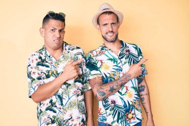 Yaz şapkası ve Hawaii tişörtü giyen iki genç eşcinsel çift endişeli ve gergin bir şekilde iki elinle, endişeli ve şaşırmış bir ifadeyi işaret ediyor. 