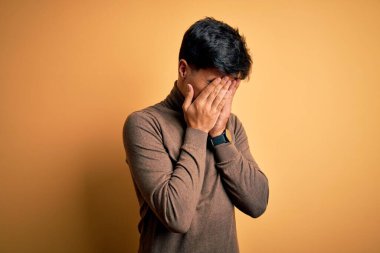 Sarı arka plan üzerine balıkçı yaka kazak giyen genç yakışıklı bir adam. Ağlarken suratını ellerle kapatan üzgün bir ifade. Depresyon kavramı.